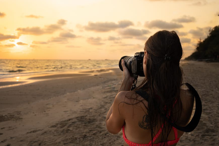 En ung kvinna riktar en SLR-kamera mot en solnedgång över en tropisk strand
