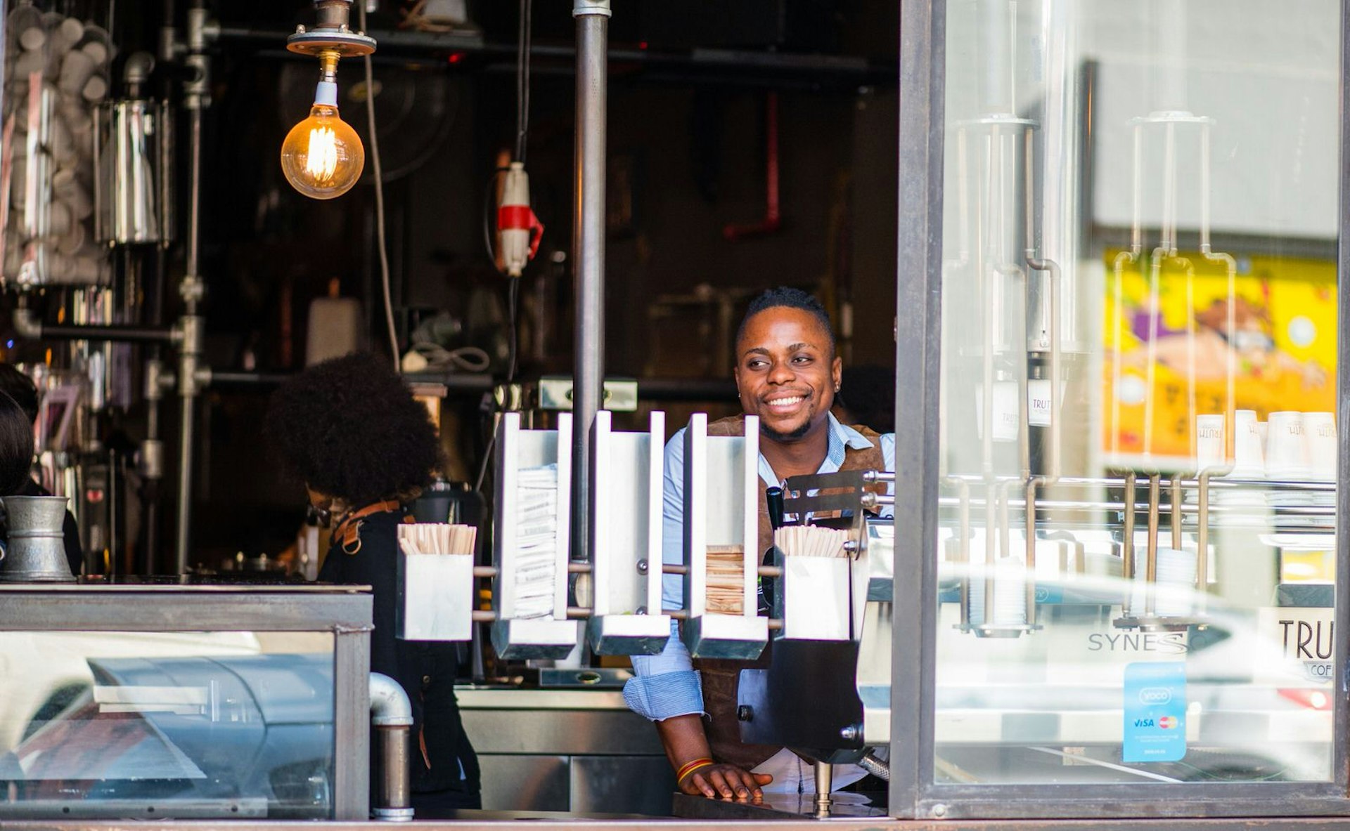 A smiling barista looks out a huge open window with a broad smile; behind him is the steampunk interior full of pipes and industrial pieces - it's an iconic scene in Cape Town coffee