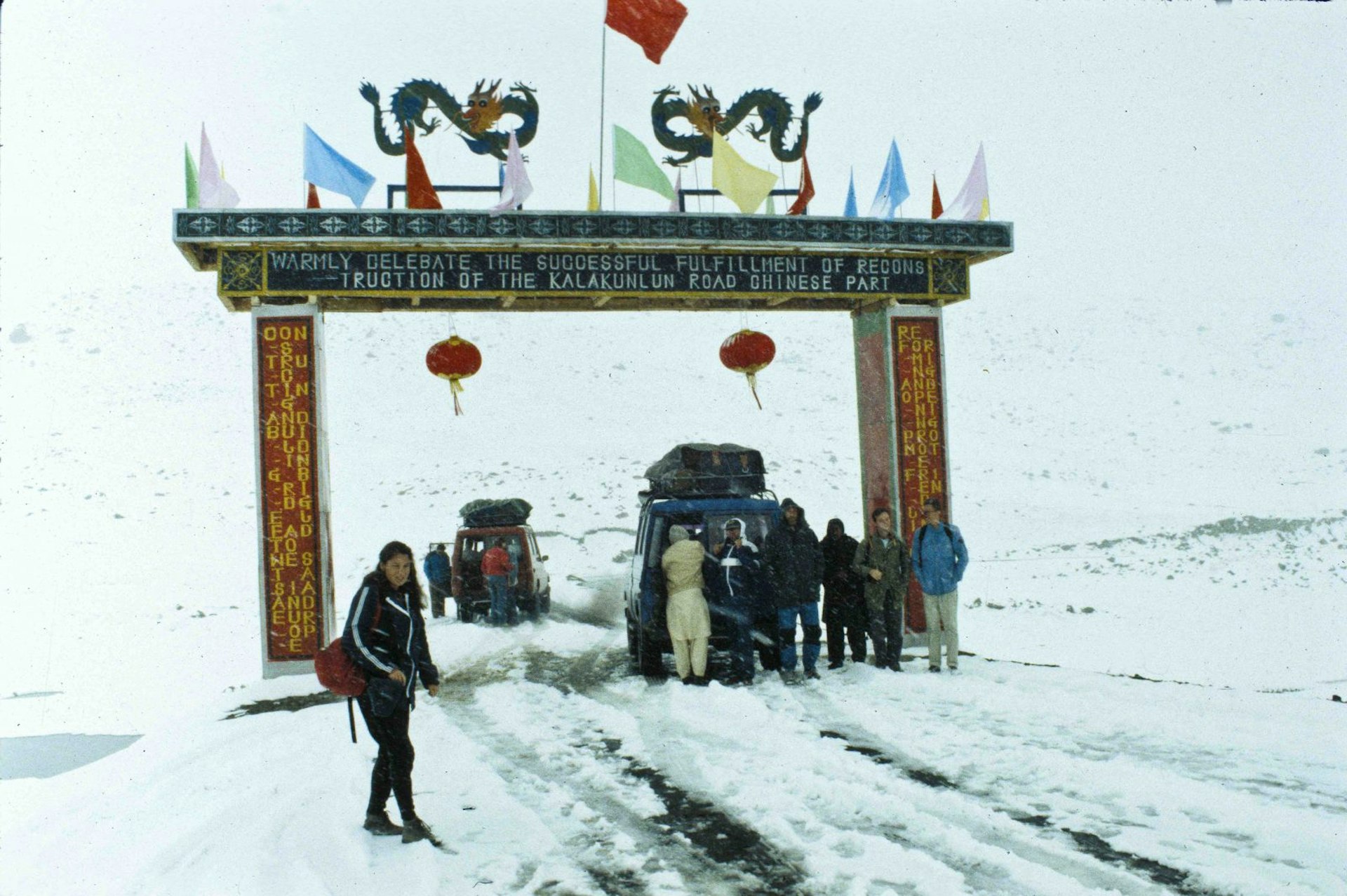 A woman stands in front of the Khunjerab Pass. An ornately decorated entrance straddles the snow-covered road. Two cars are behind her, one stopped under the entrance and one just the far side of it. People are standing around both vehicles in heavy clothing.
