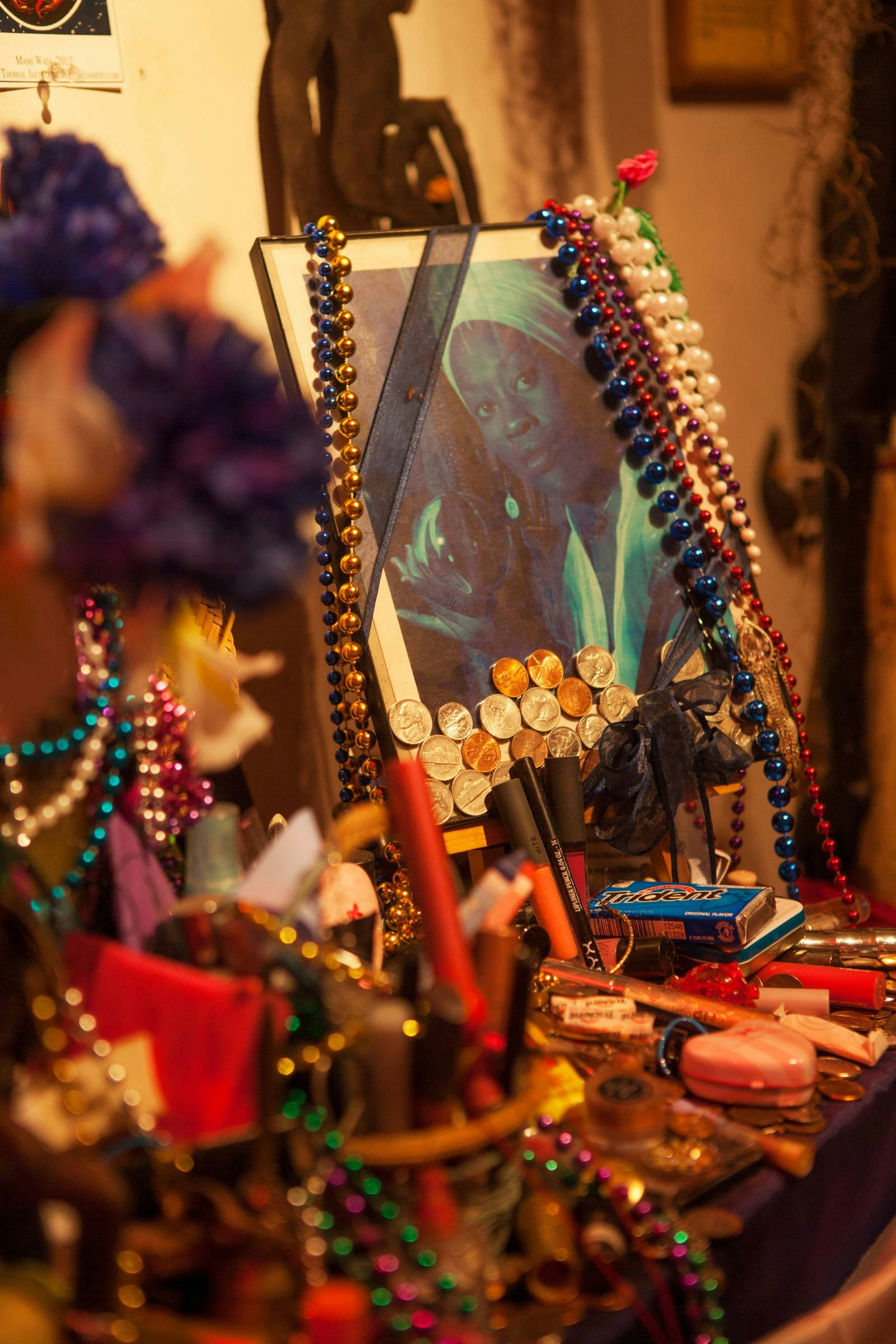 A framed picture stands in the middle of an altar with a photo of a woman with long fingernails in a white turban, clothes and earrings holding a crystal ball. Strings of beads surround it, along with coins, gum packets, pens and a heart-shaped box.