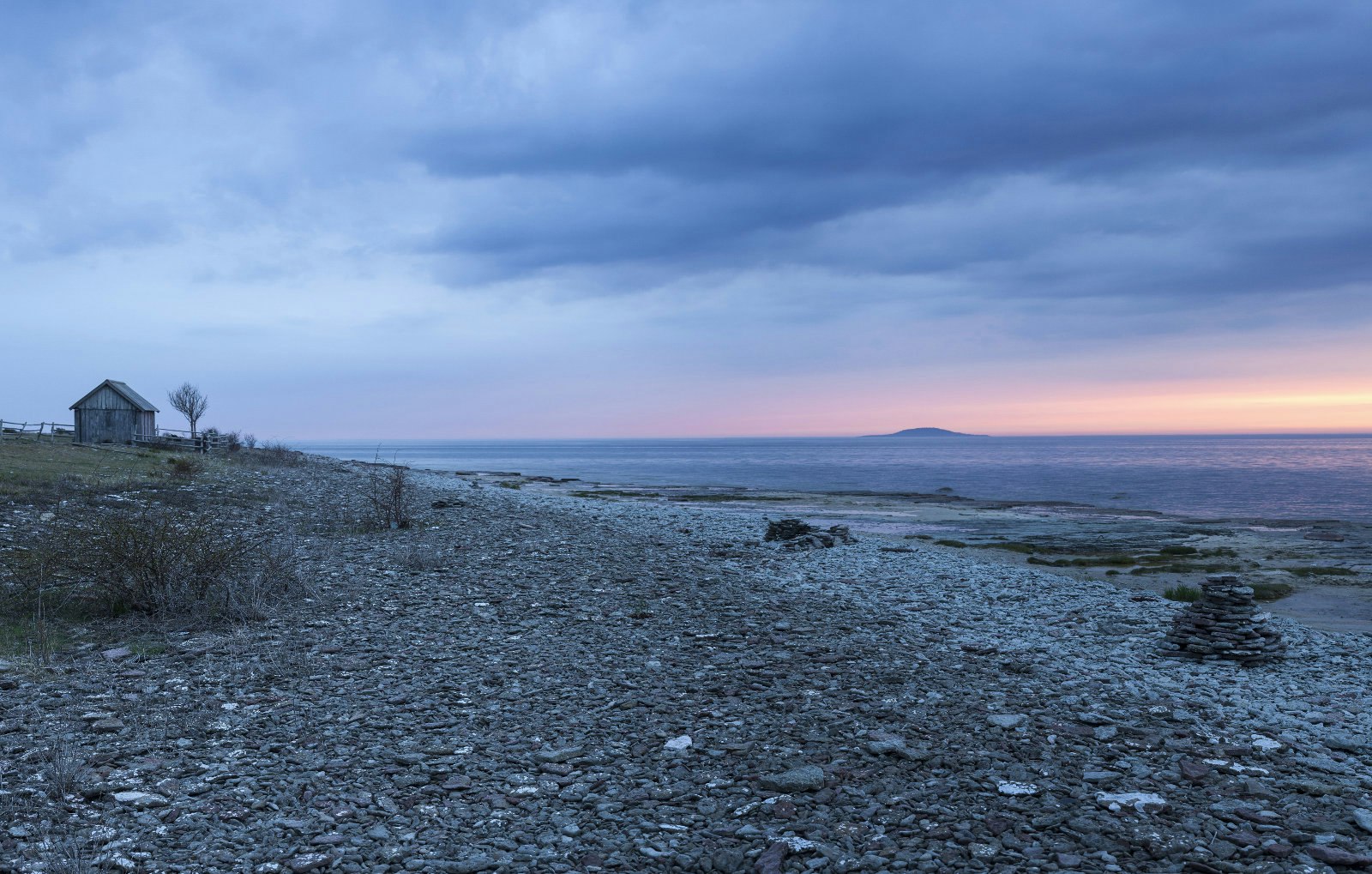A stony beach after sunset with a sliver of pink in the sky. On the left background is an old timber hut with one bare tree next to it.