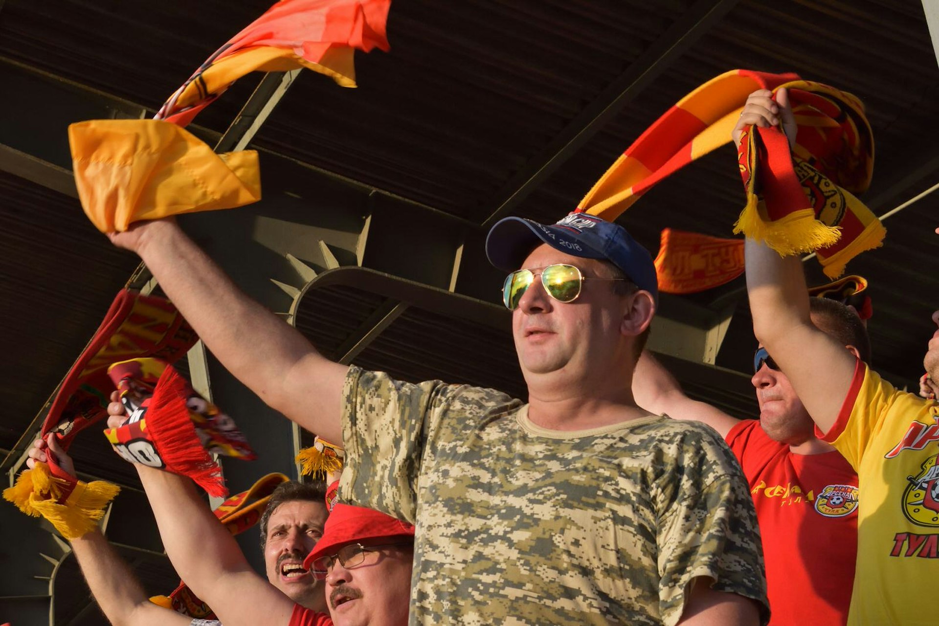A Tulsa fan waves a red and yellow scarf in the air at a football match. The man is wearing a green, camouflage-print t-shirt, baseball cap and aviator sunglasses. 