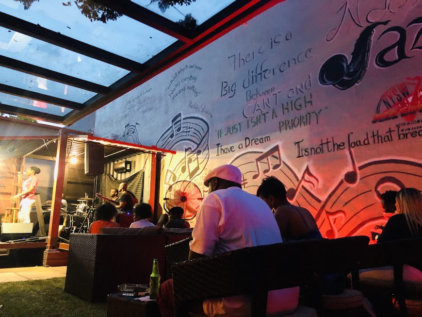 A side view of a jazz singer on the stage at NYSW Jazz Lounge. A perspex roof covers the stage and seating area next to it as patrons drink and enjoy the music. The wall next to them is covered in graffiti. 