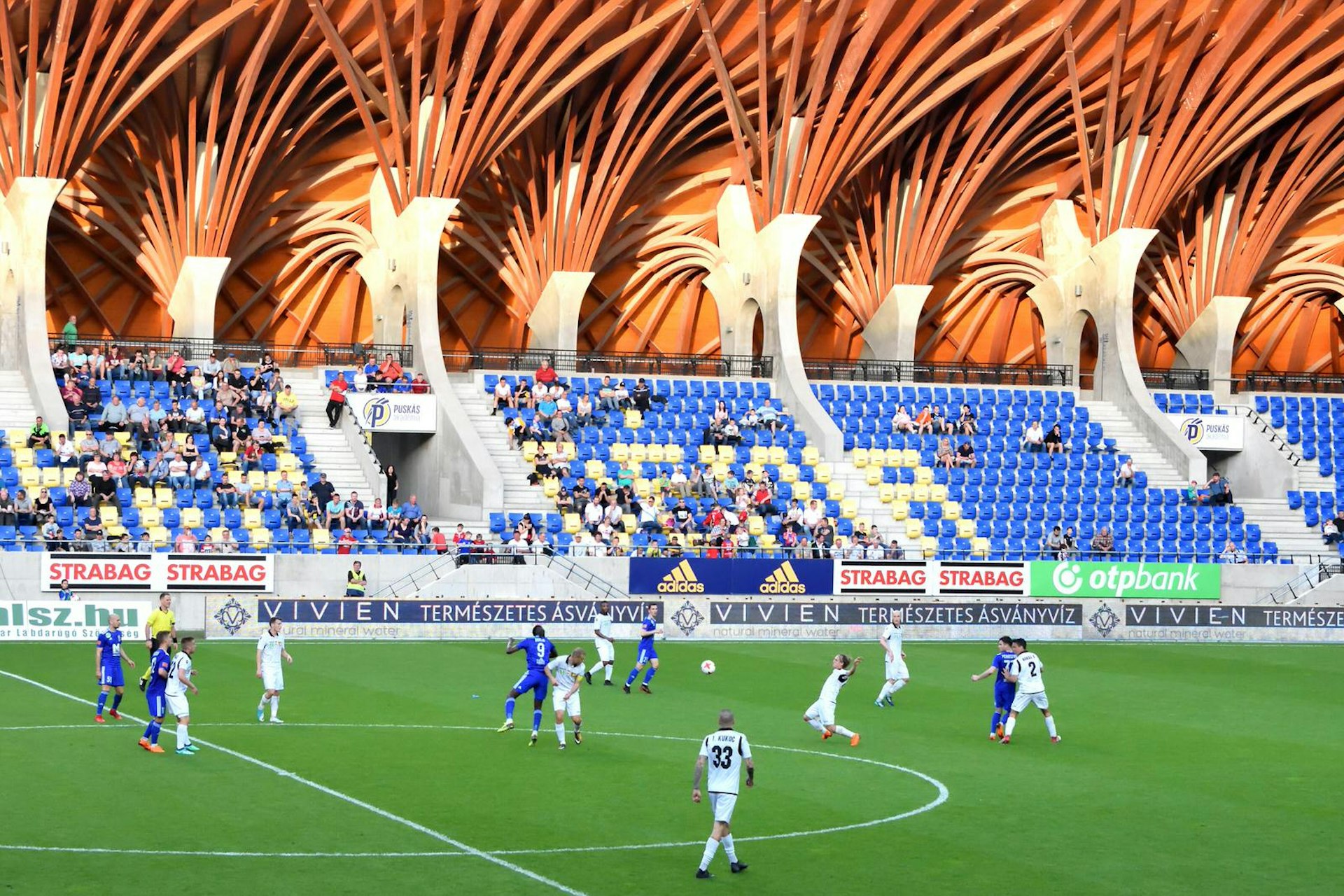 A football match in action at the Pancho Aréna. Beyond the pitch a stand of spectators is visible and above them is the sweeping, timber-beamed roof that makes the Pancho Aréna so distinctive.