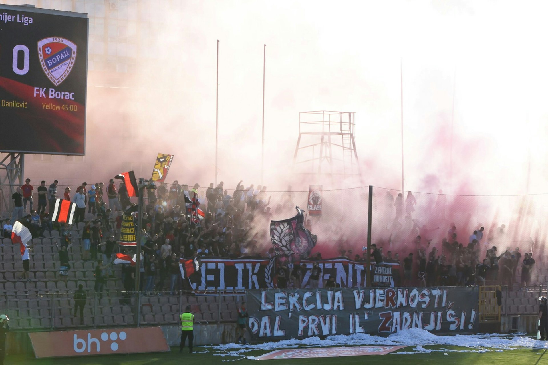 Groups of football fans in a stand during a Čelik home game. Flares have been let off, with the smoke obscuring many of the people in the crowd.