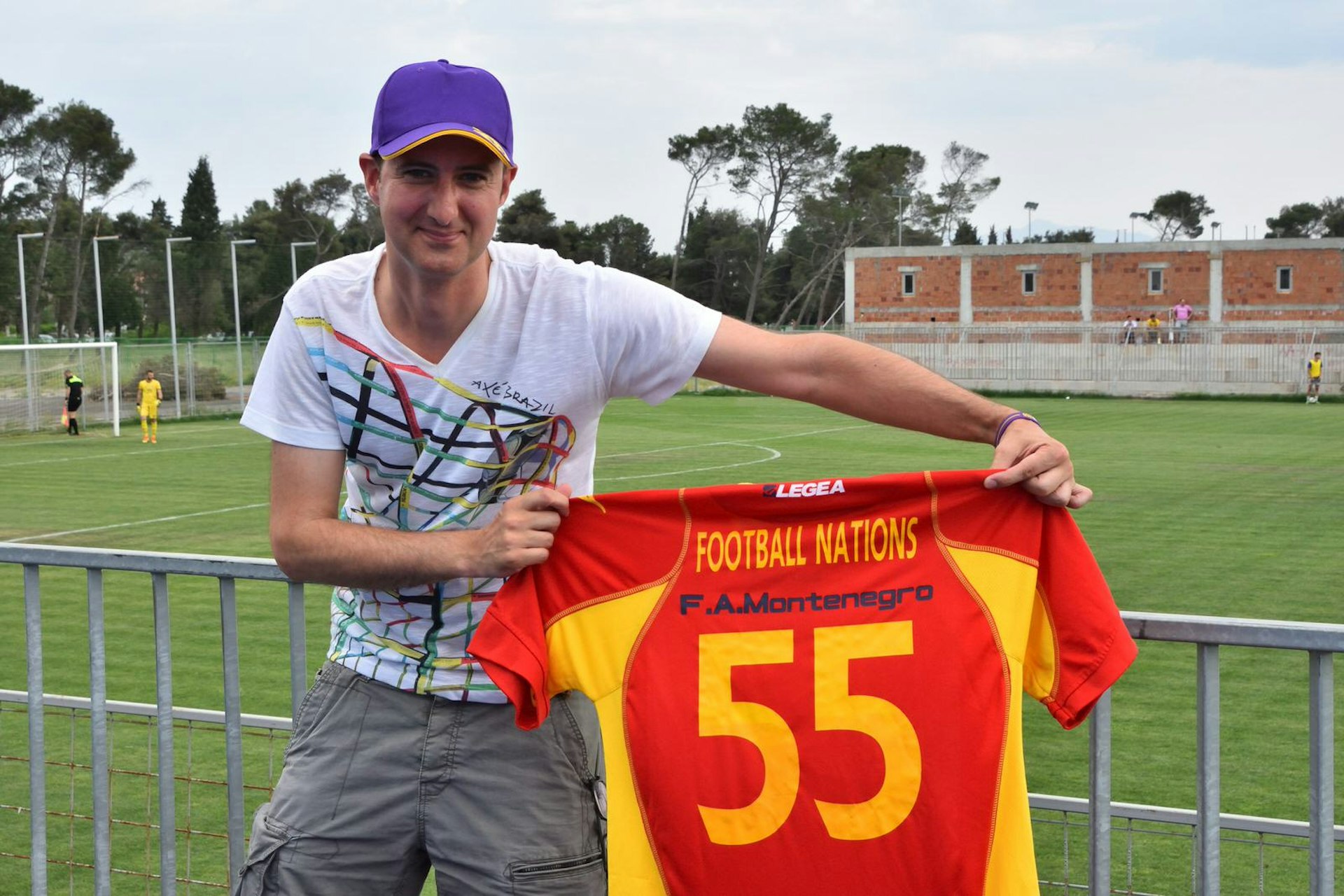 Matt stands next to a football pitch in Montenegro, holding a shirt with the number 55 on it