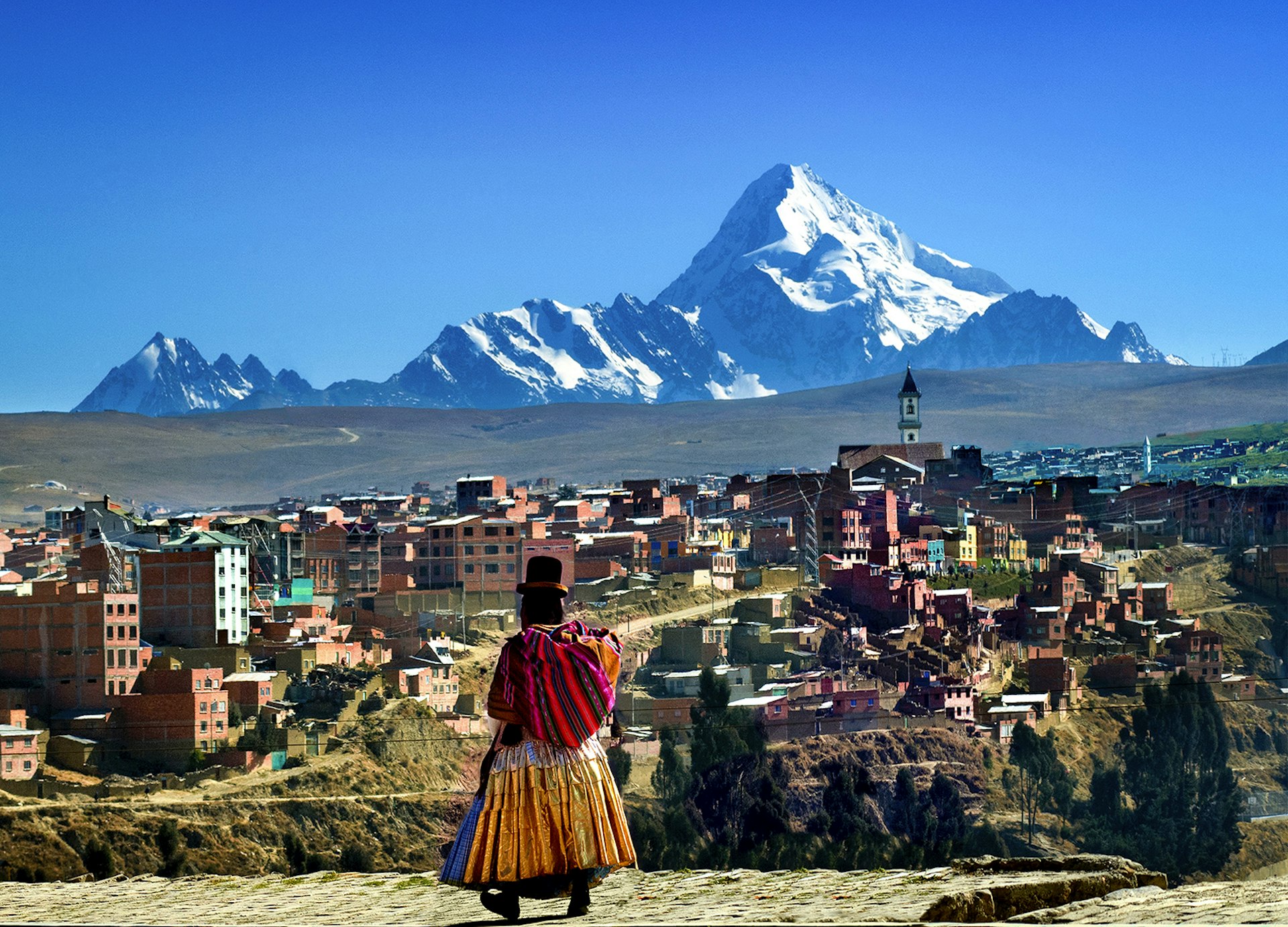 A woman in traditional Bolivian dress walks on a high ridge in La Paz, with the city and a snowcapped mountain in the background. Bolivia.