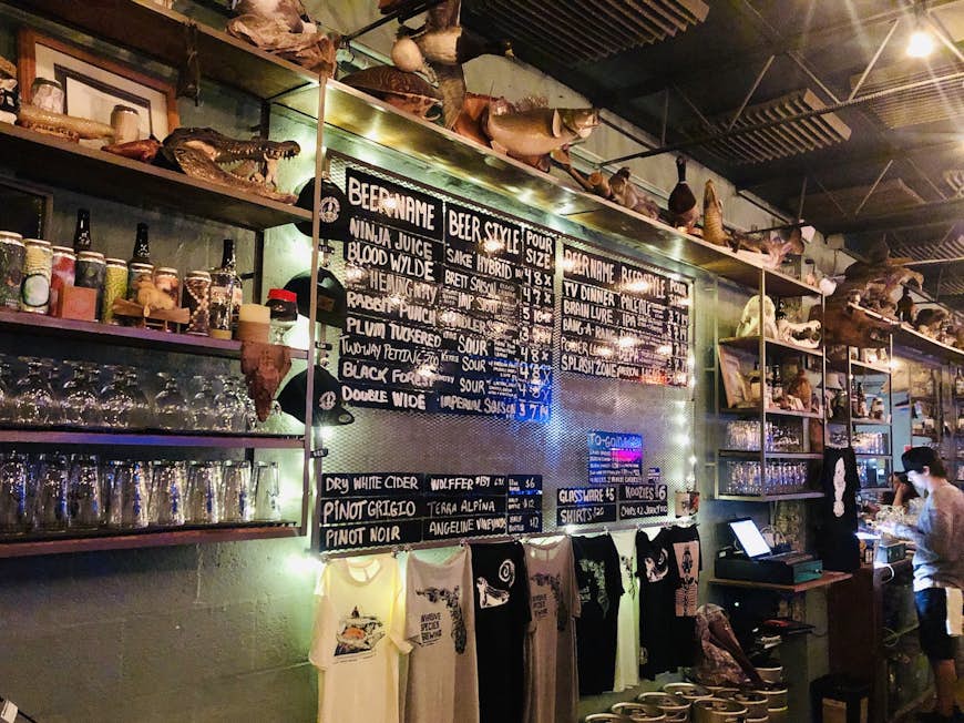 An interior shot of the bar with a wall covered in shelves of liquor, glasses, t-shirts, a beer menu and various items of taxidermy.