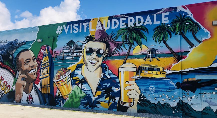 A colorful mural adorns a large wall. It depicts a man with a British flag tie with Big Ben next to him on the left and a man in a Hawaiian shirt and holding a beer is in the centre of the image. Various other images of Fort Lauderdale are included.
