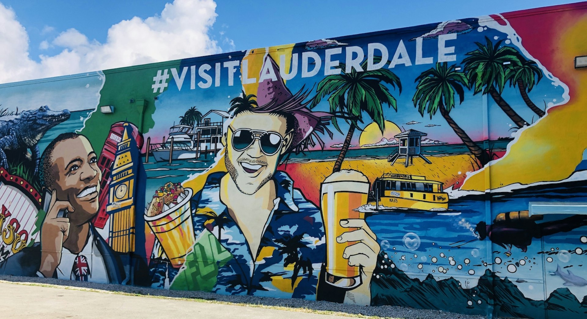 A colorful mural adorns a large wall. It depicts a man with a British flag tie with Big Ben next to him on the left and a man in a Hawaiian shirt and holding a beer is in the centre of the image. Various other images of Fort Lauderdale are included.