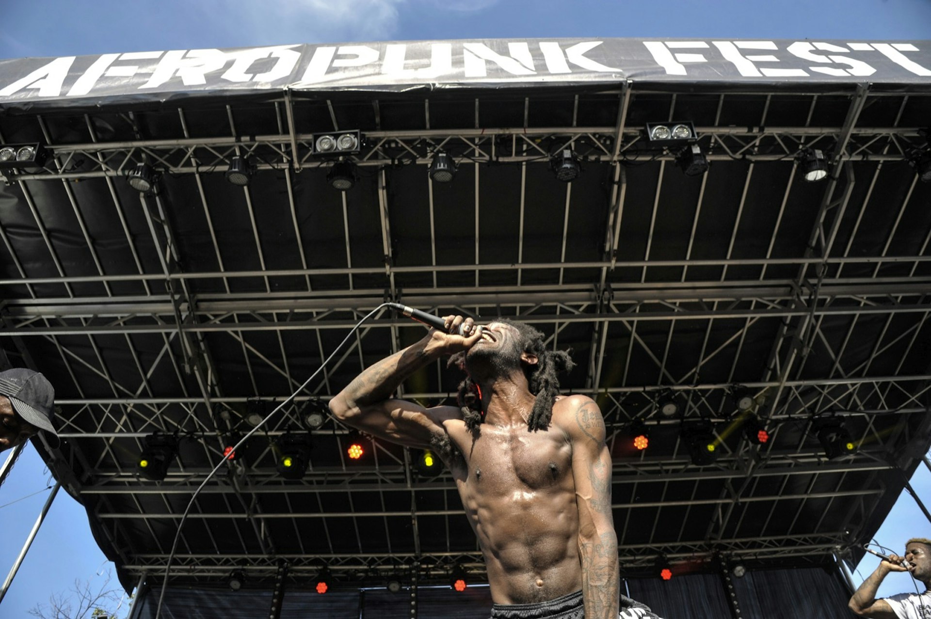 Photo taken from below a shirtless performer who screams into a microphone, leaning over the edge of a stage. Above him is a sign that says Afropunk fest. 