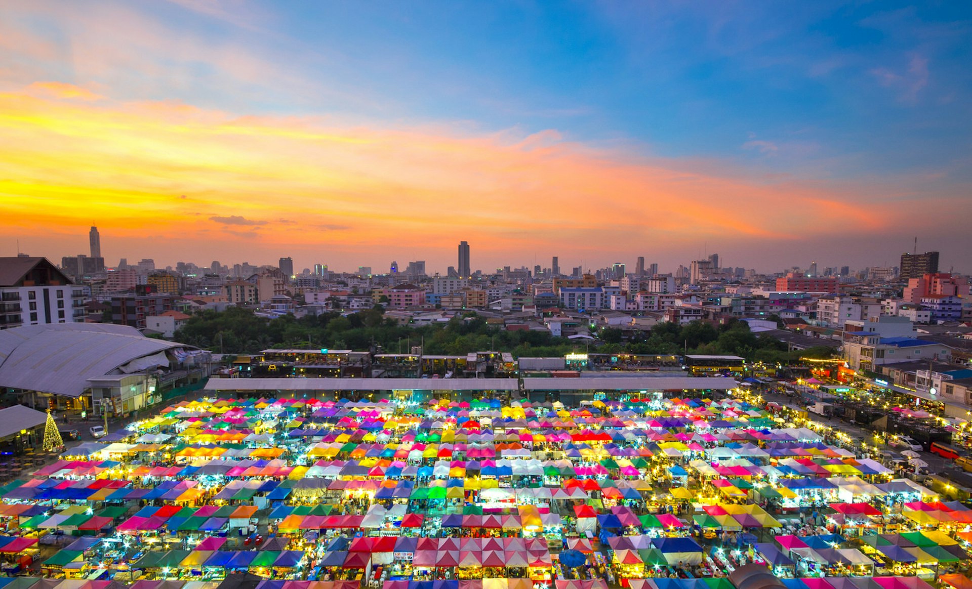 Hemmed in by a dark cityscape beneath a golden sunset is a brilliant square of vibrantly coloured stalls at the Chatuchak Weekend Market