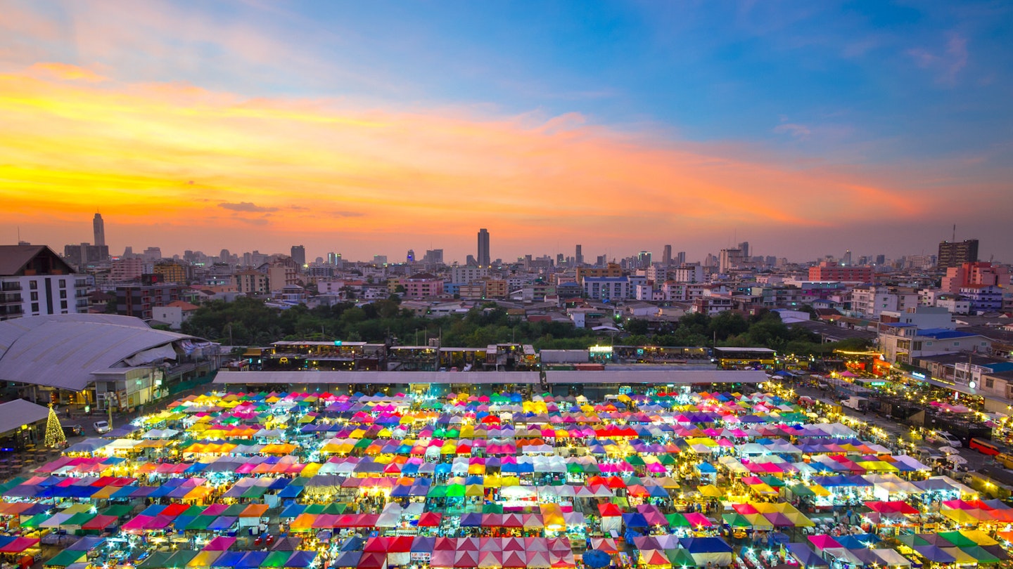 Hemmed in by a dark cityscape beneath a golden sunset is a brilliant square of vibrantly coloured stalls at the Chatuchak Weekend Market