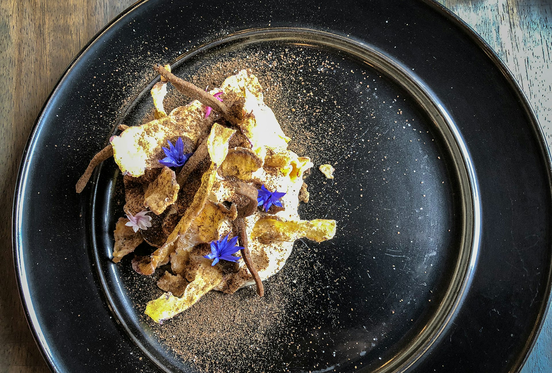 A close-up of modern Bolivian food on a black plate, sprinkled with brown powder and garnished with small purple and pink flowers. La Paz, Bolivia.