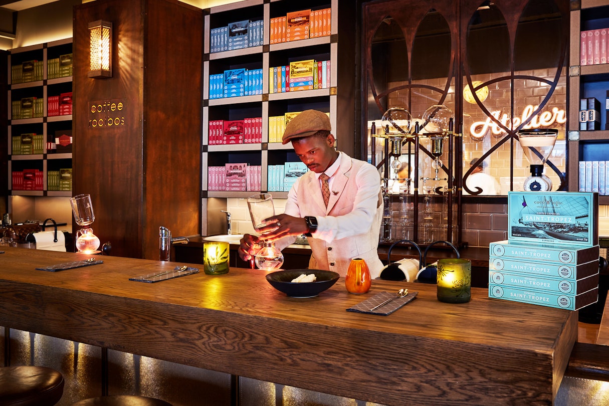 A smartly dressed barista in a flat cap, waist coat and tie concocts a coffee; the rich wood interior includes numerous shelves full of boxed coffee (each shelve holding matching coloured boxes)