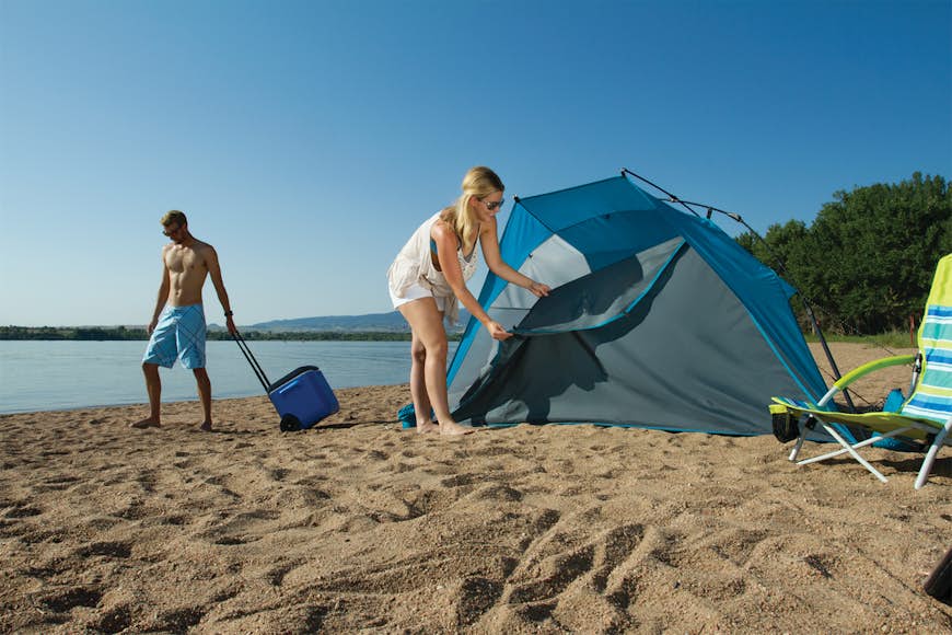 En kvinna reser ett tält på en strand medan en man utan bar överkropp stirrar i marken med en kylare;  strandutrustning