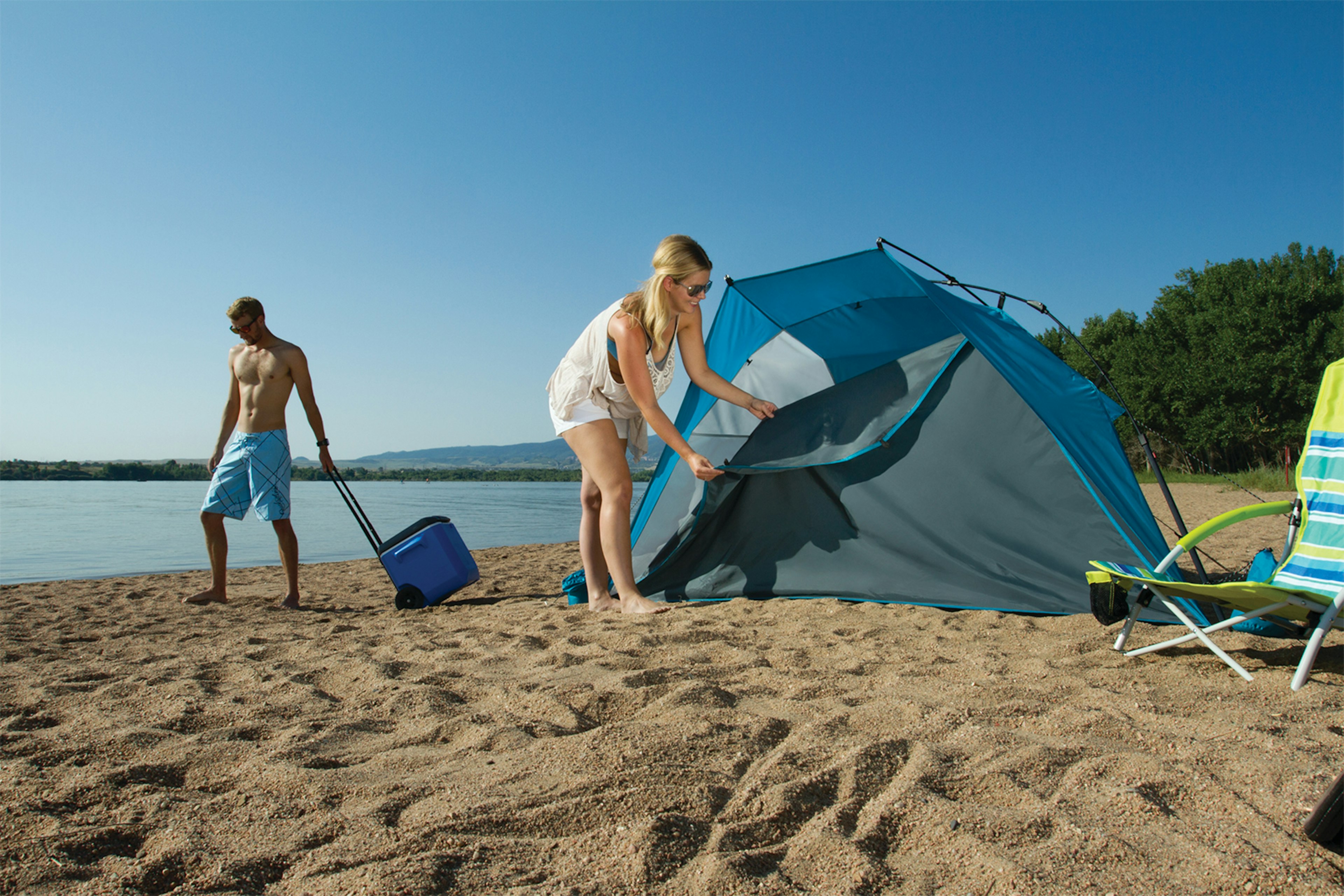 A woman erects a tent on a beach while a shirtless man poses with a cooler