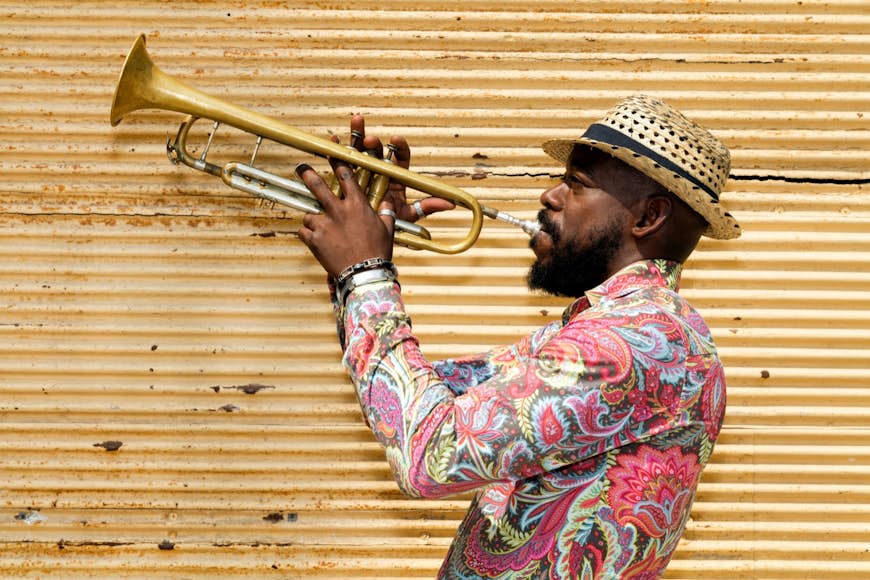 Cuban musician with a beard, wearing a Panama hat and colorful shirt standing against a closed yellow store shutter, playing a trumpet outdoors; Cuba