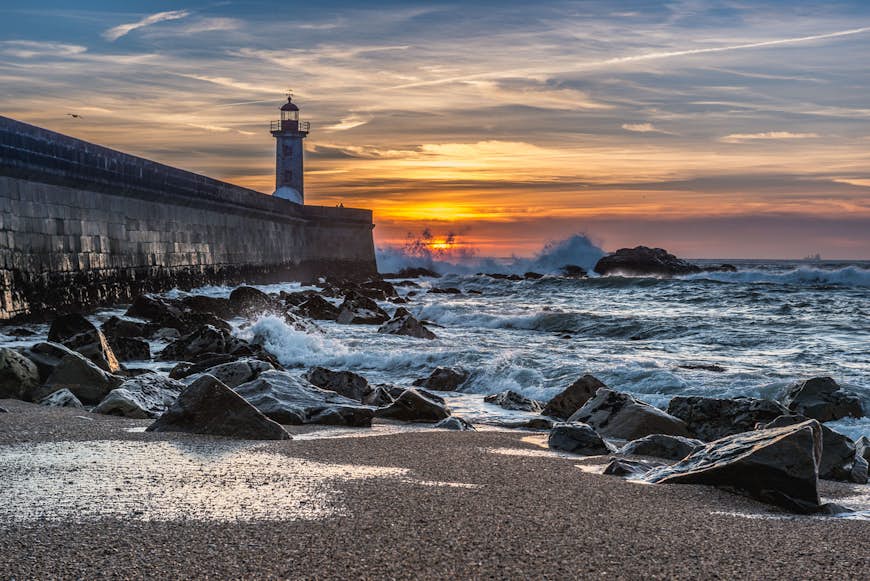 Felgueiras Lighthouse stands at the end of a concrete pier; in the distance the sun sets into a rough ocean, while in the foreground the sea rolls over a beach covered in large stones; free things to do in Porto