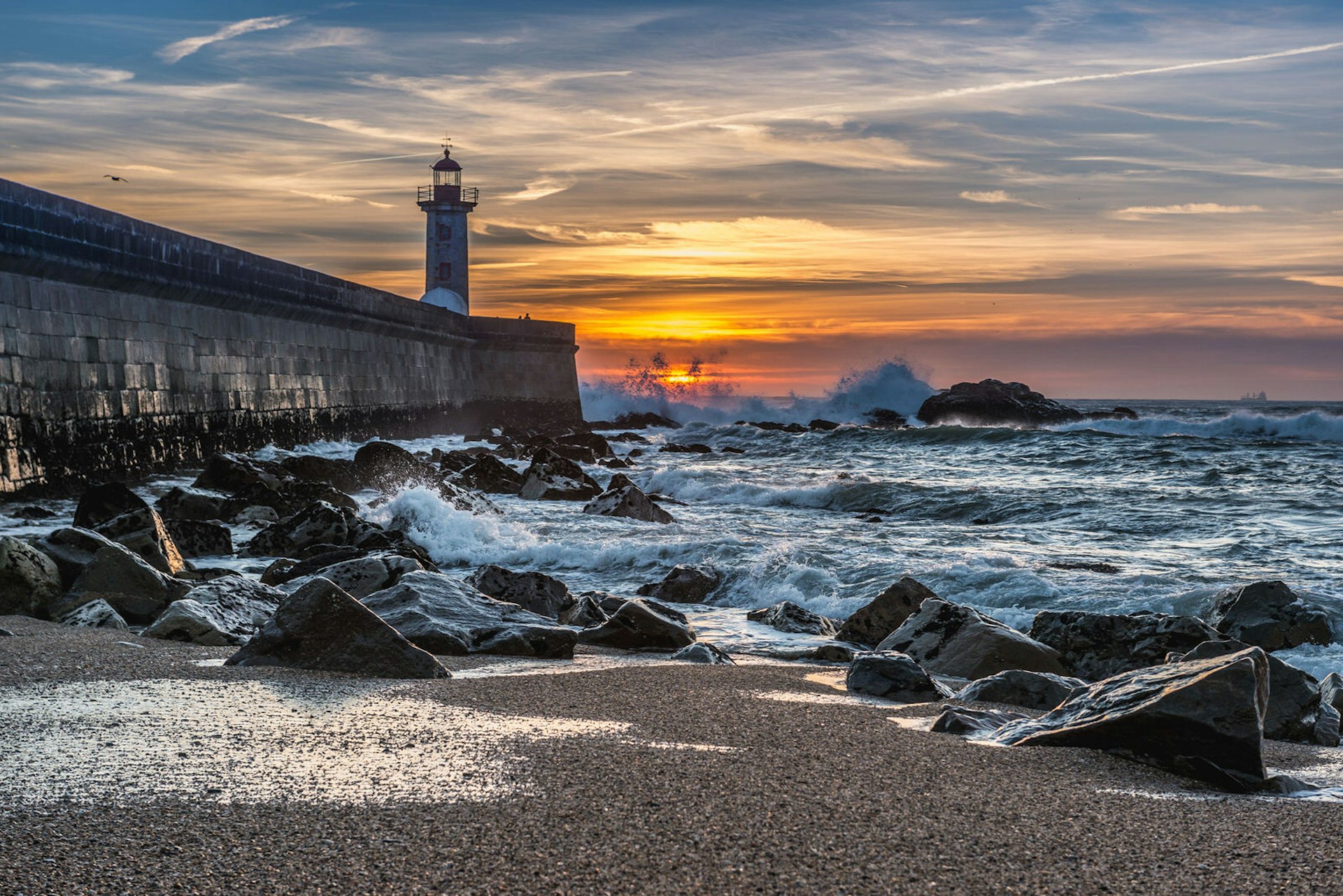 Felgueiras Lighthouse stands at the end of a concrete pier; in the distance the sun sets into a rough ocean, while in the foreground the sea rolls over a beach covered in large stones; free things to do in Porto