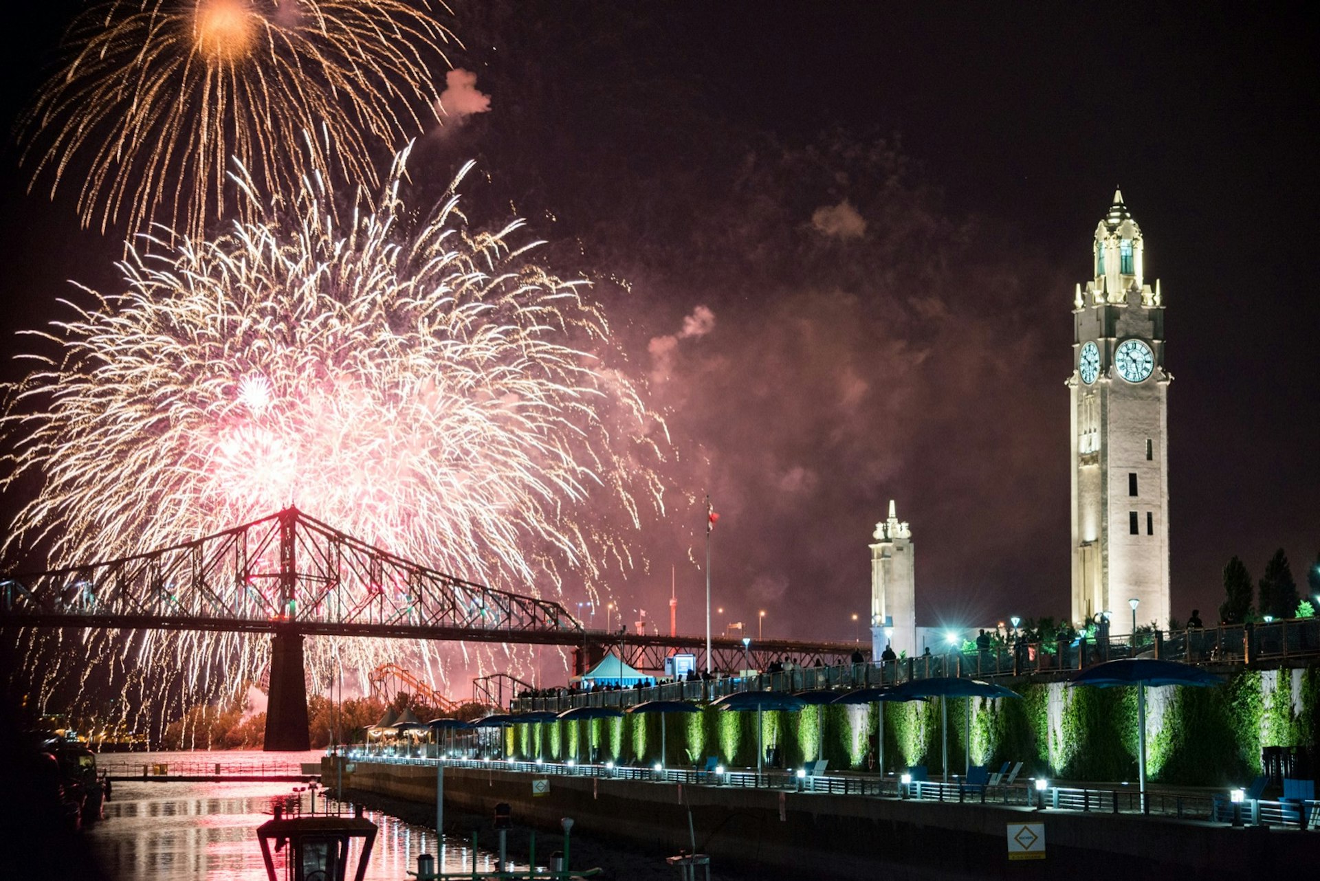 Fireworks explode in the sky over a bridge and iconic clock tower. The International Fireworks Festival is one of the best free things to do in Montréal
