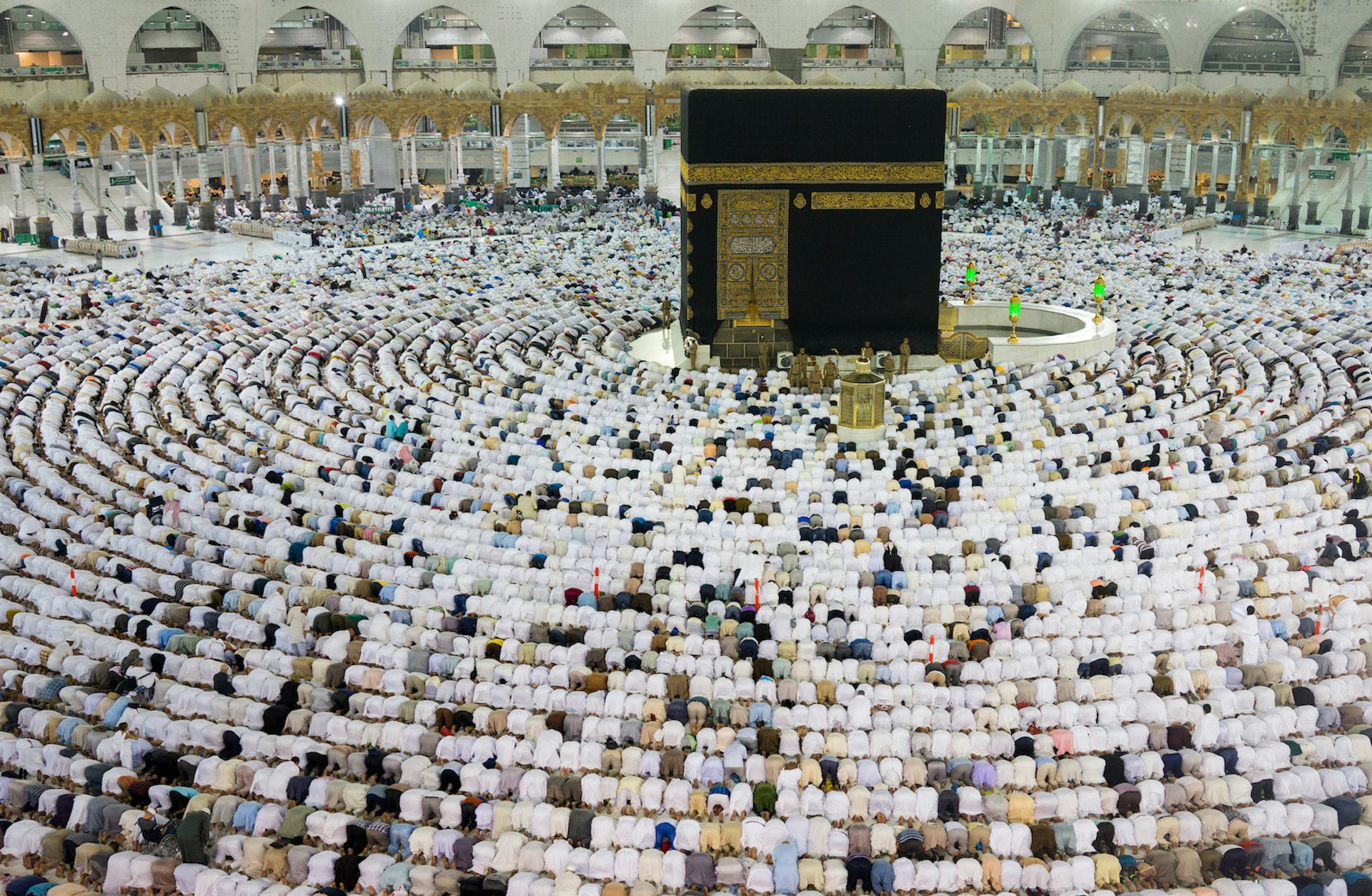 Thousands of Muslims are kneeling in concentric circles around the Kaaba at the The Great Mosque in Mecca. The Kaaba is a large black stone cube with ornate gold details. The majority of the worshippers are wearing white. 