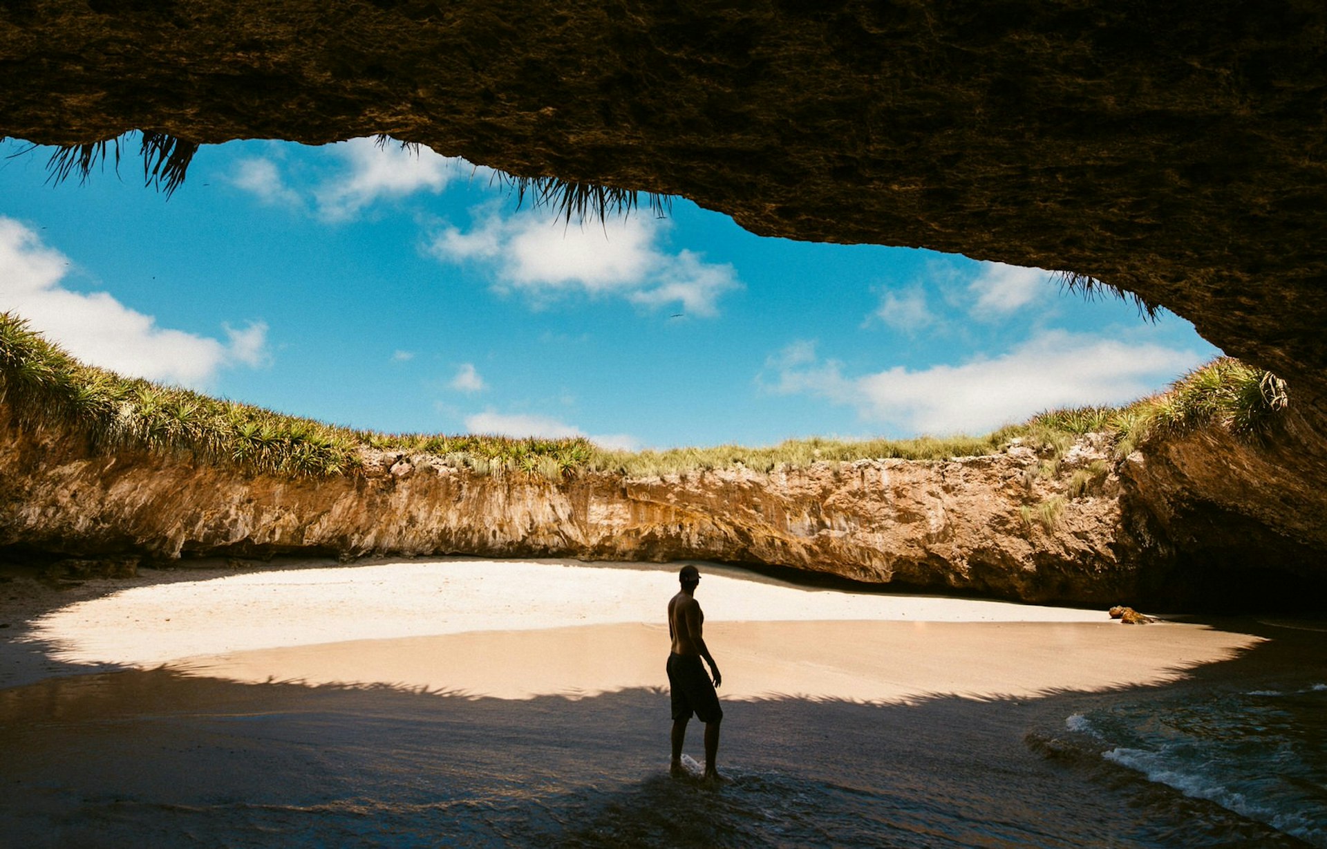 La Playa del Amor: a man stands on the beach that is in a circular cavern and surrounded by rock on all sides. Above a chunk of blue sky with a smattering of fluffy clouds is visible.