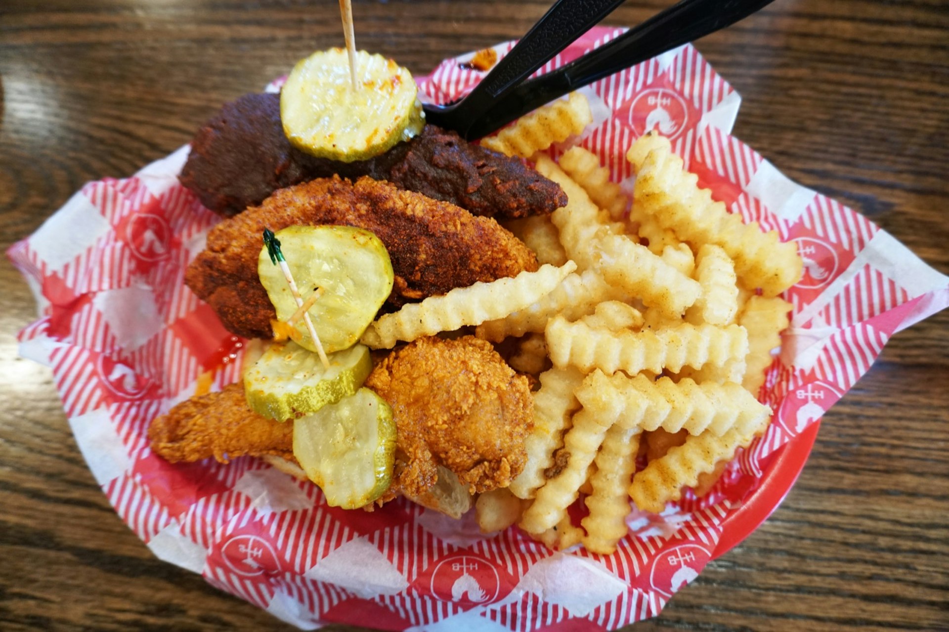 Ett foto av varma kycklinganbud knivhuggna med tandpetare och pickles bredvid skrynkliga pommes frites ovanpå rött och vitt papper i en röd skål;  helgen Nashville 