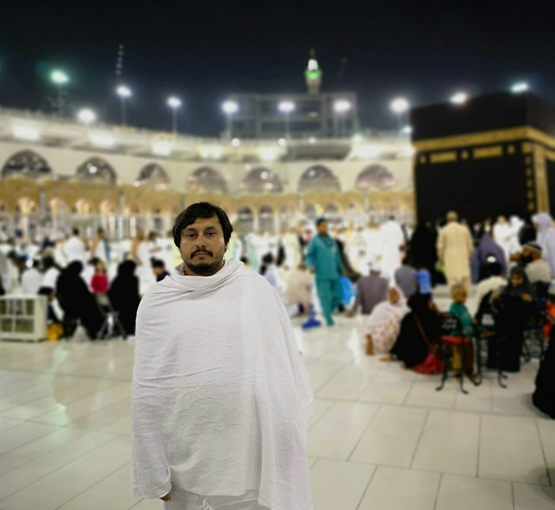 A man in white robe stands facing the camera in front of the Kaaba in The Great Mosque in Mecca. A large group of pilgrims are in the background behind him, sitting, standing and talking.