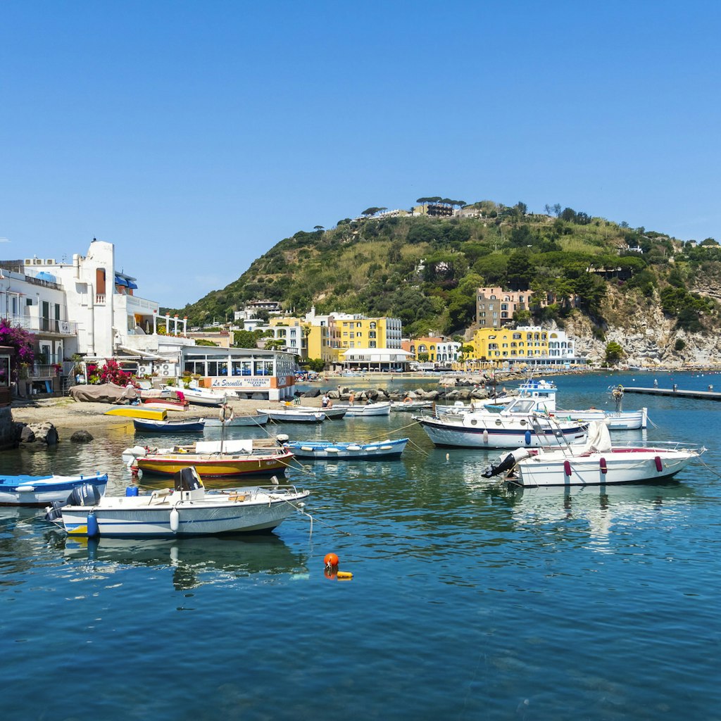 Several small boats moored along the coast of Ischia. There are restaurants and hotels in the background, nestled among hills and mediterranean foliage.