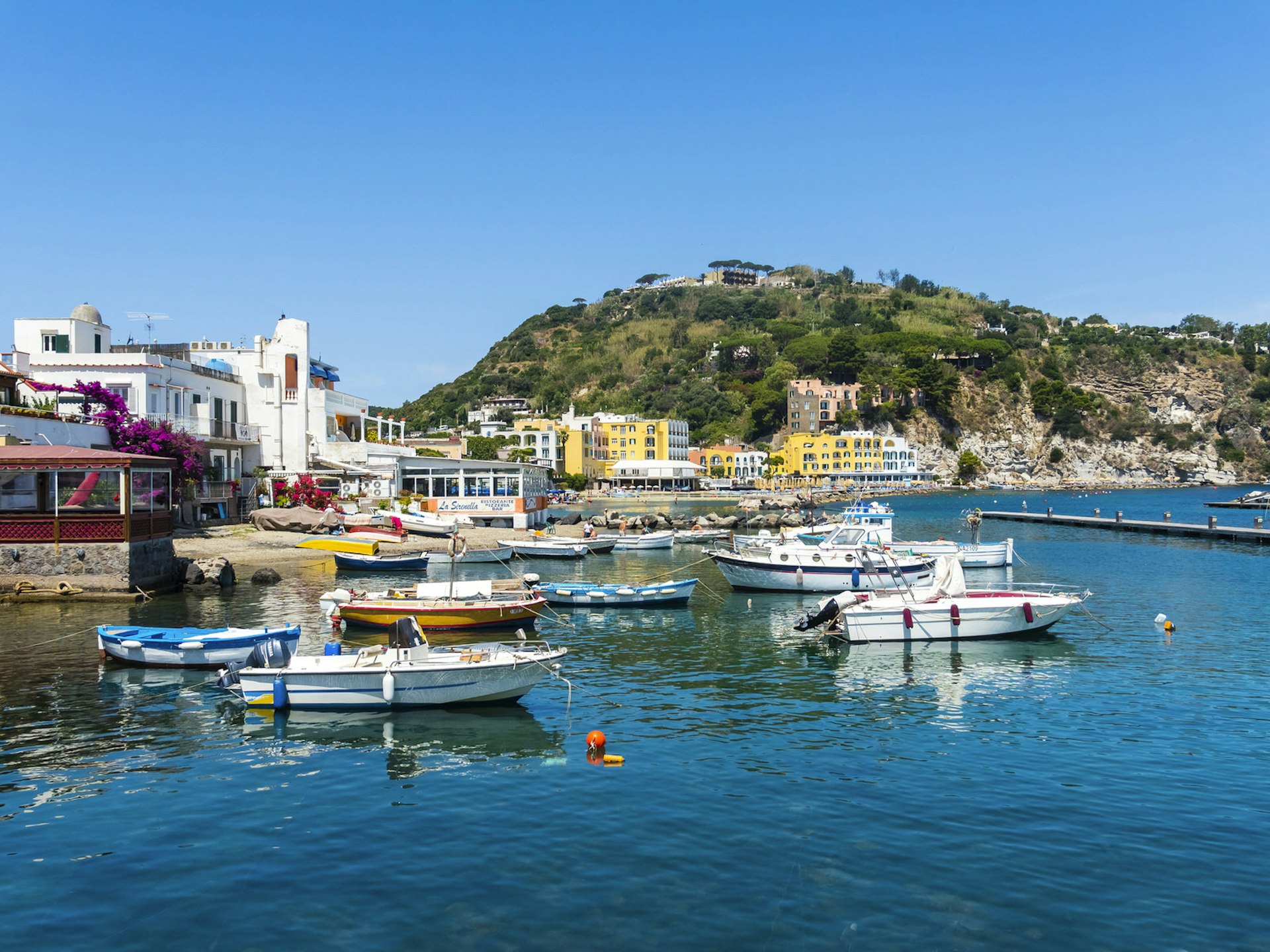 Several small boats moored along the coast of Ischia. There are restaurants and hotels in the background, nestled among hills and Mediterranean foliage.