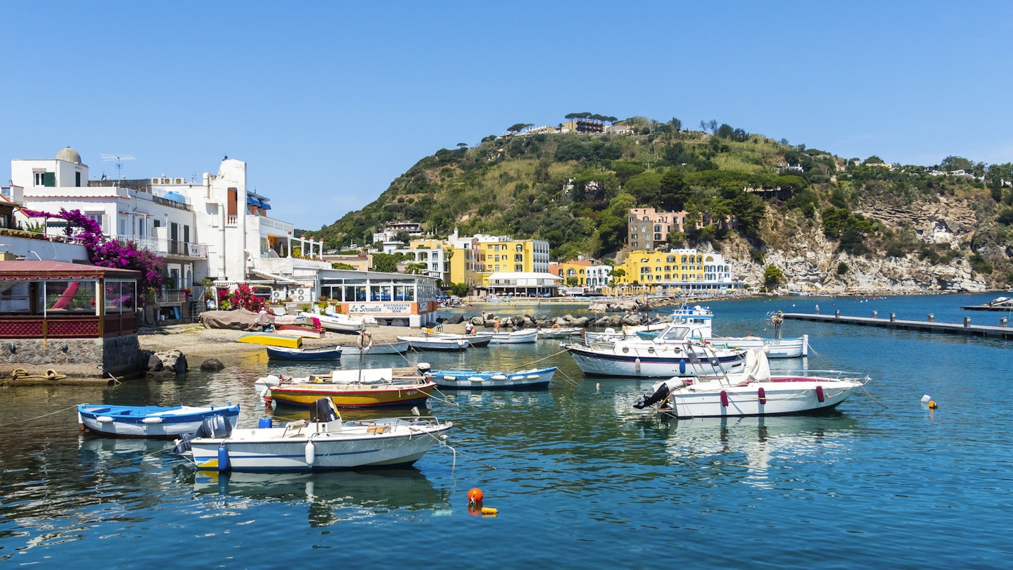 Several small boats moored along the coast of Ischia. There are restaurants and hotels in the background, nestled among hills and mediterranean foliage.