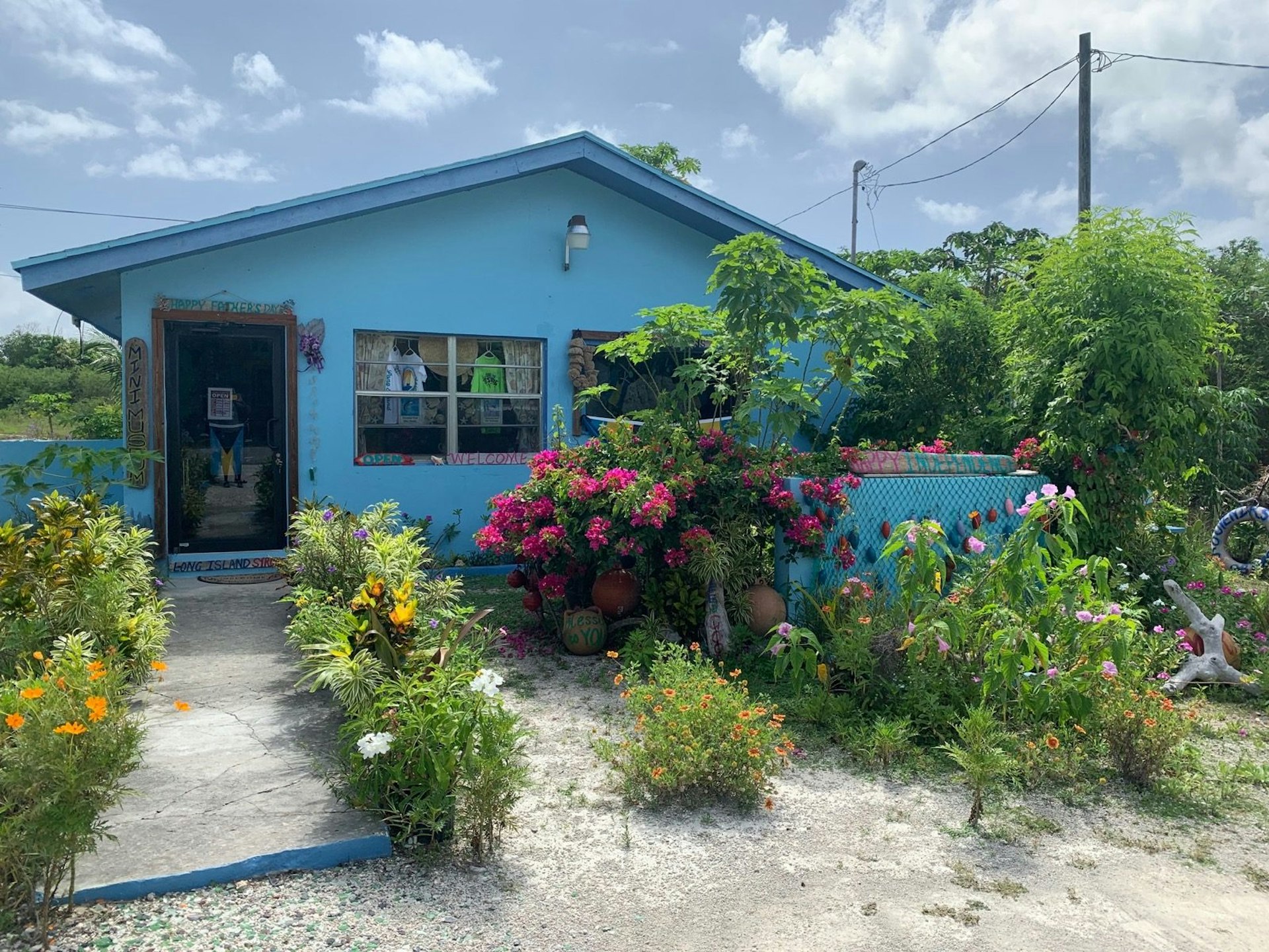 A large garden filled with colorful bright pink and yellow flowers in front of a bright blue house; Long Island Bahamas 