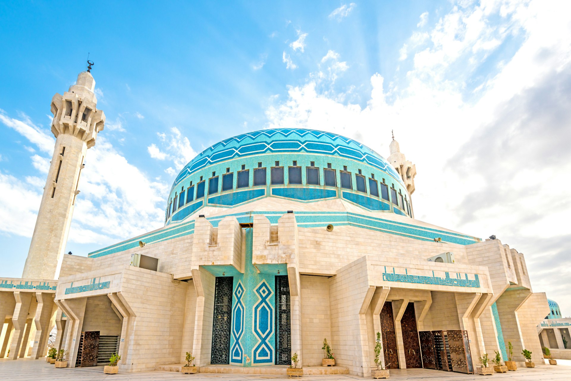 Shot with a wide-angle lens, this image looks from the ground up at the large blue dome and towering minarets; the lower part of the mosque is tan coloured with blue mosaic tiles; Free things to do in Amman, Jordan