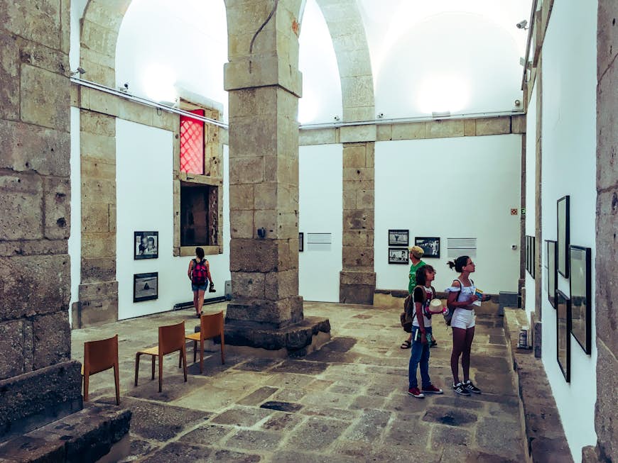 Fyra personer tittar på olika fotoutställningar i ett stort vitkalkat rum i ett gammalt fängelse;  stora stenpelare och valv dominerar utrymmet;  gratis saker att göra i Porto