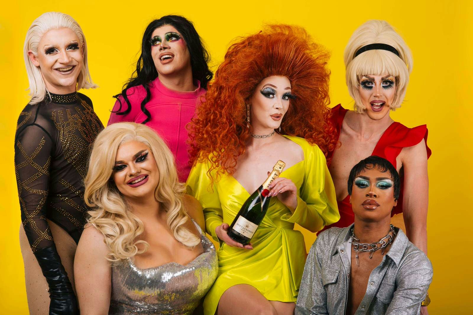 A group of drag queens pose in front of a solid yellow background. The queen in the centre wears a yellow dress and huge red-haired wig, holding a bottle of prosecco. London drag brunches