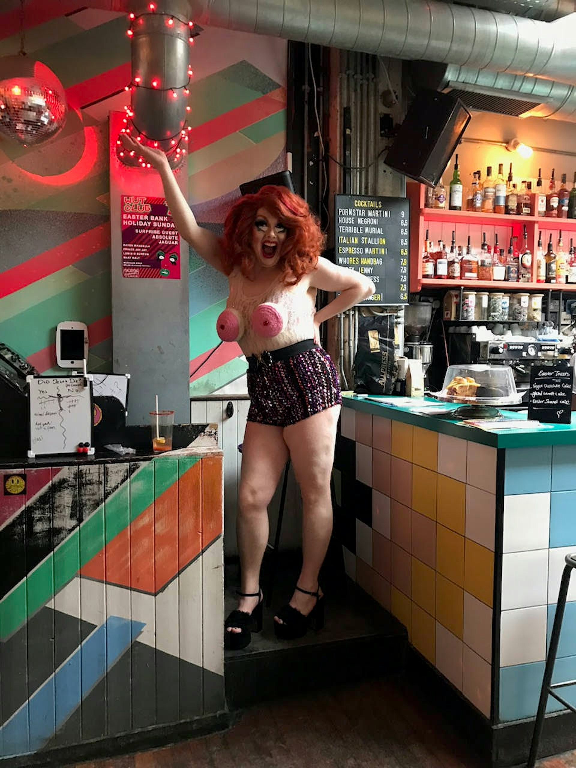 En dragqueen ställer sig i en stickad topp med rosa bröst och stickade hotpants samt full make up och en rödhårig peruk.  Dalston Superstore har en bardel med färgglada kakel och neonljus på väggen.  London dragbruncher