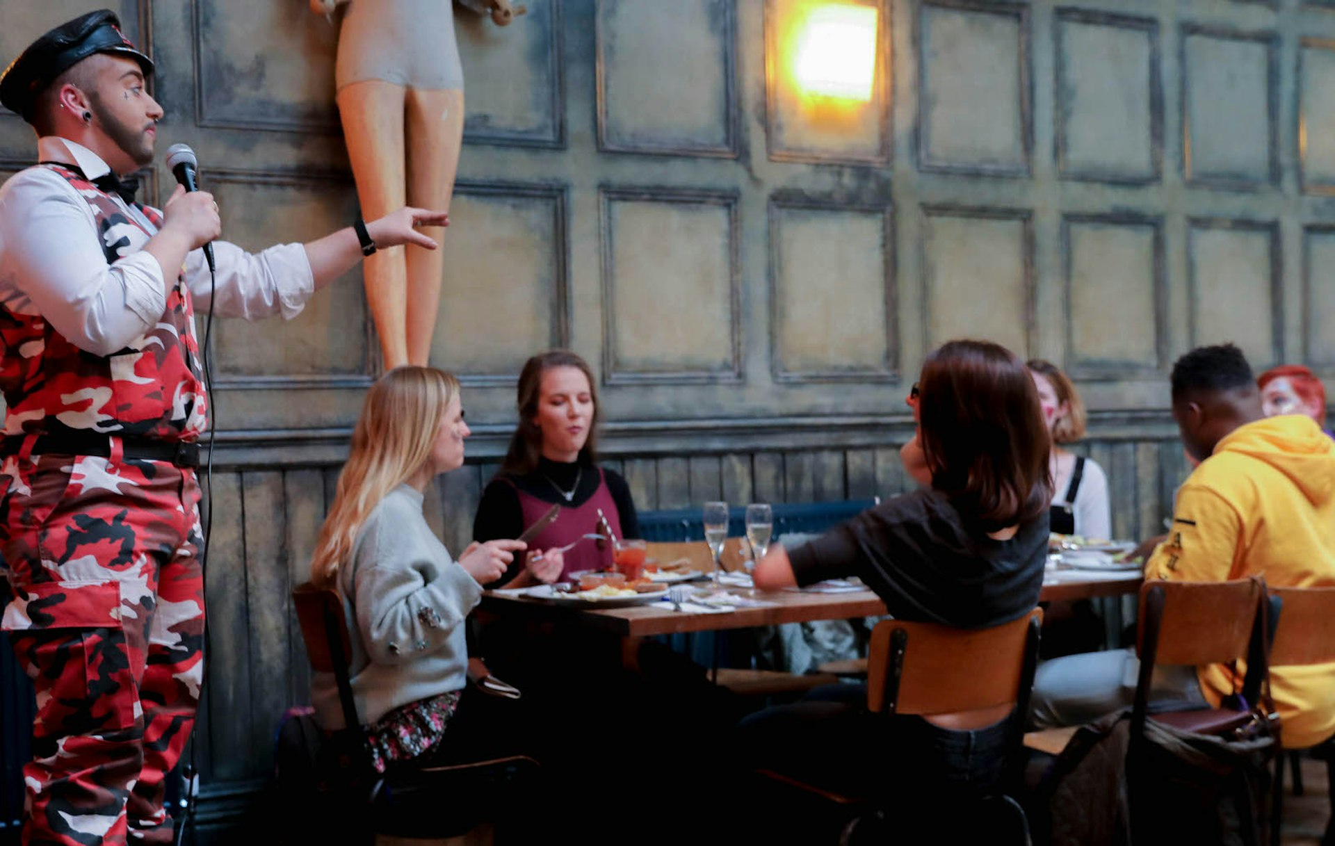 A group of people sit around a table eating brunch and laughing while a drag queen hosts the entertainment wearing pink camouflage overalls, pointing somewhere off camera. There is a blow-up doll on the wall. London drag brunches