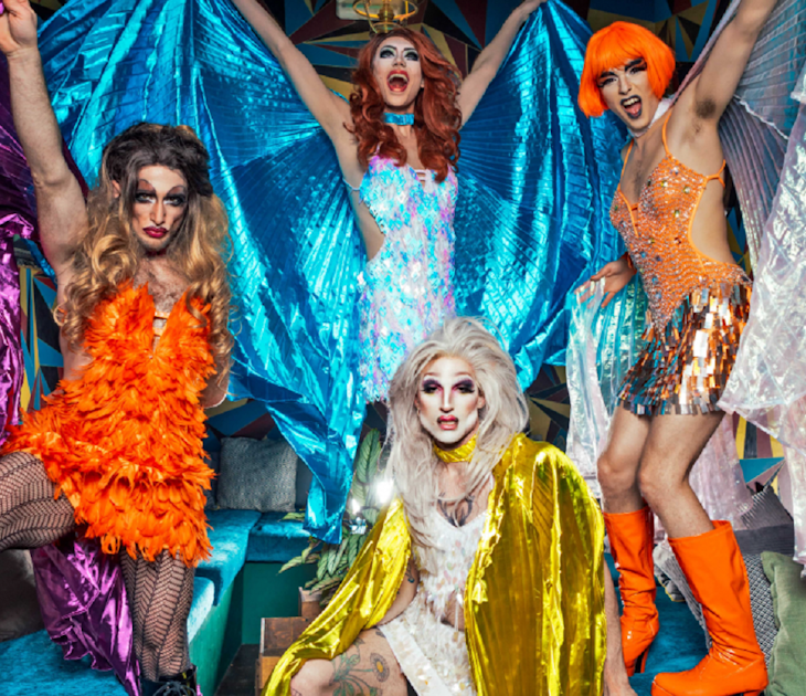 Four drag queen pose on a a table. They are wearing metallic and feathery costumes in orange, blue, white and yellow.