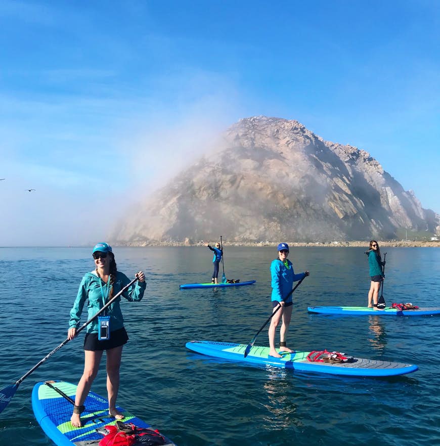 Morro Rock stands out in Morro Bay, covered in fog, as a group of paddleboarders in blue jackets explores the water around it