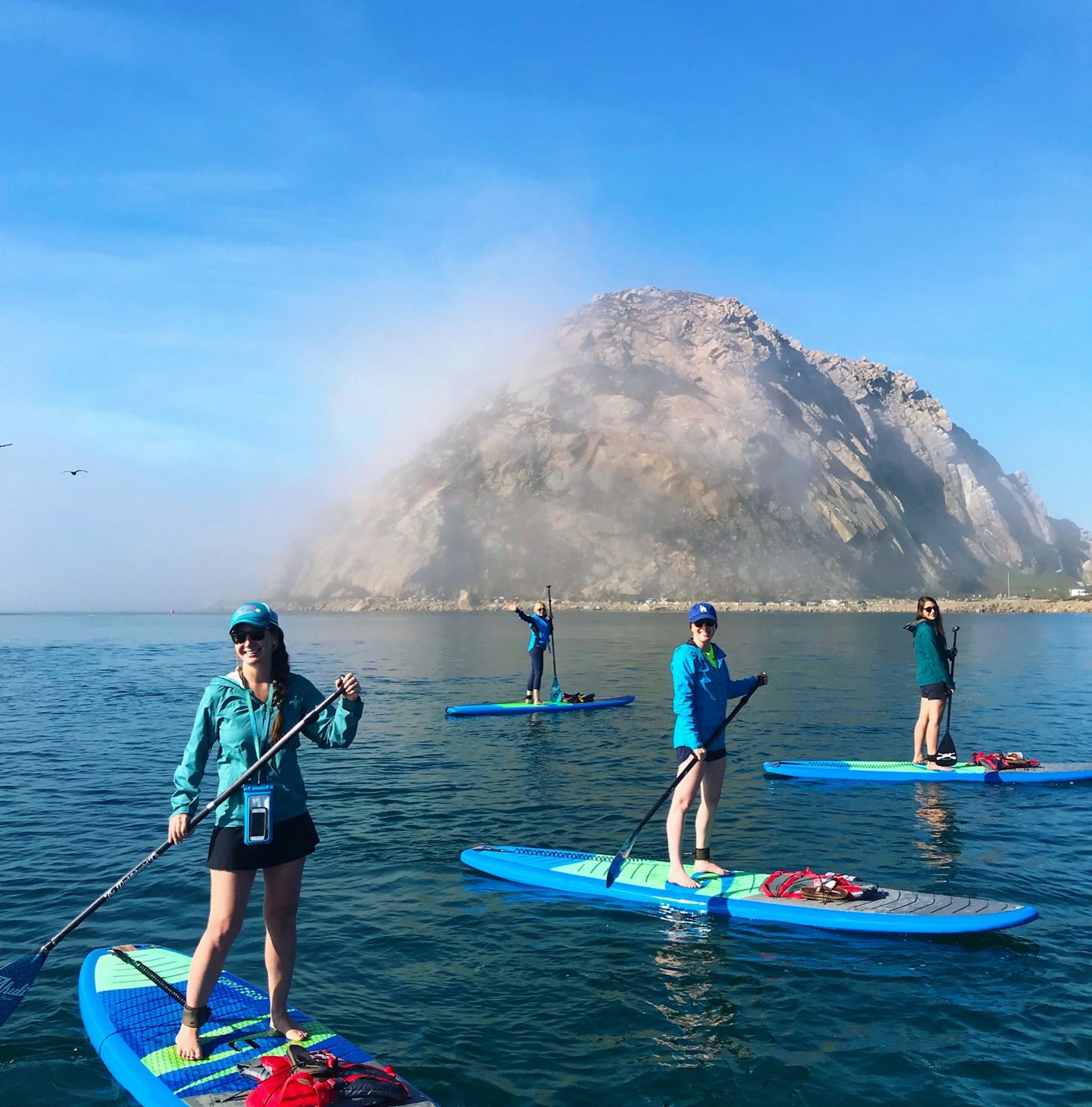Morro Rock stands out in Morro Bay, covered in fog, as a group of paddleboarders in blue jackets explores the water around it