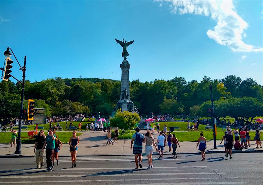 People walk across a street toward a park with a large statue in the middle topped by a winged figure; free things to do in Montréal