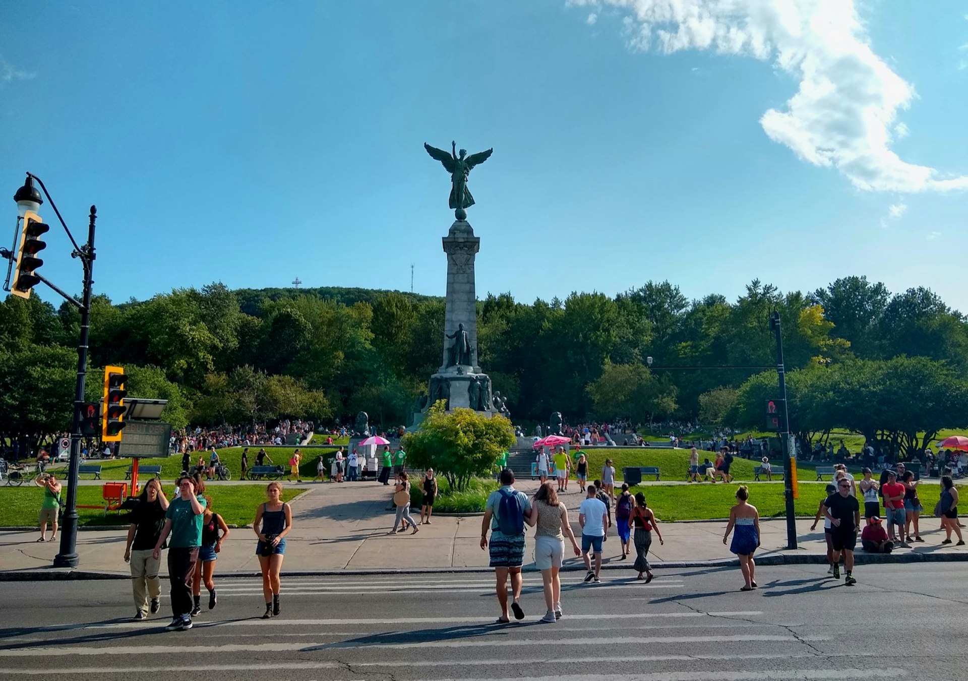 People walk across a street toward a park with a large statue in the middle topped by a winged figure; free things to do in Montréal