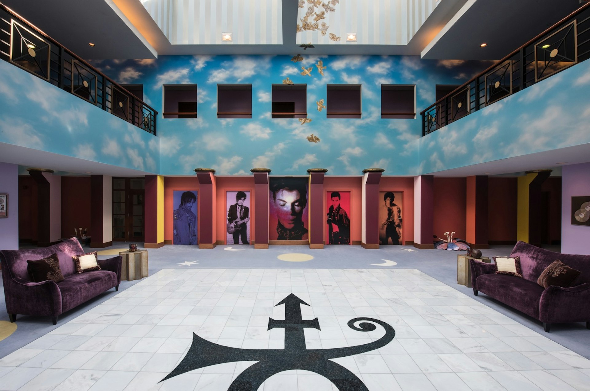 Prince's symbol is in the middle of a white floor in black, as images of the singer at various points in his career look out from cubbyholes in a sky-blue wall with clouds and two balconies on either side of the atrium