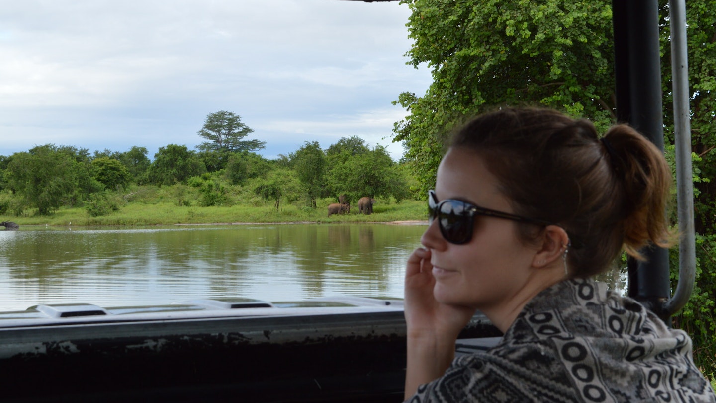 A woman wearing sunglasses sits in a jeep looking out over a lake in Udawalawe, Sri Lanka. There is a small group of elephants on the other side of the lake.