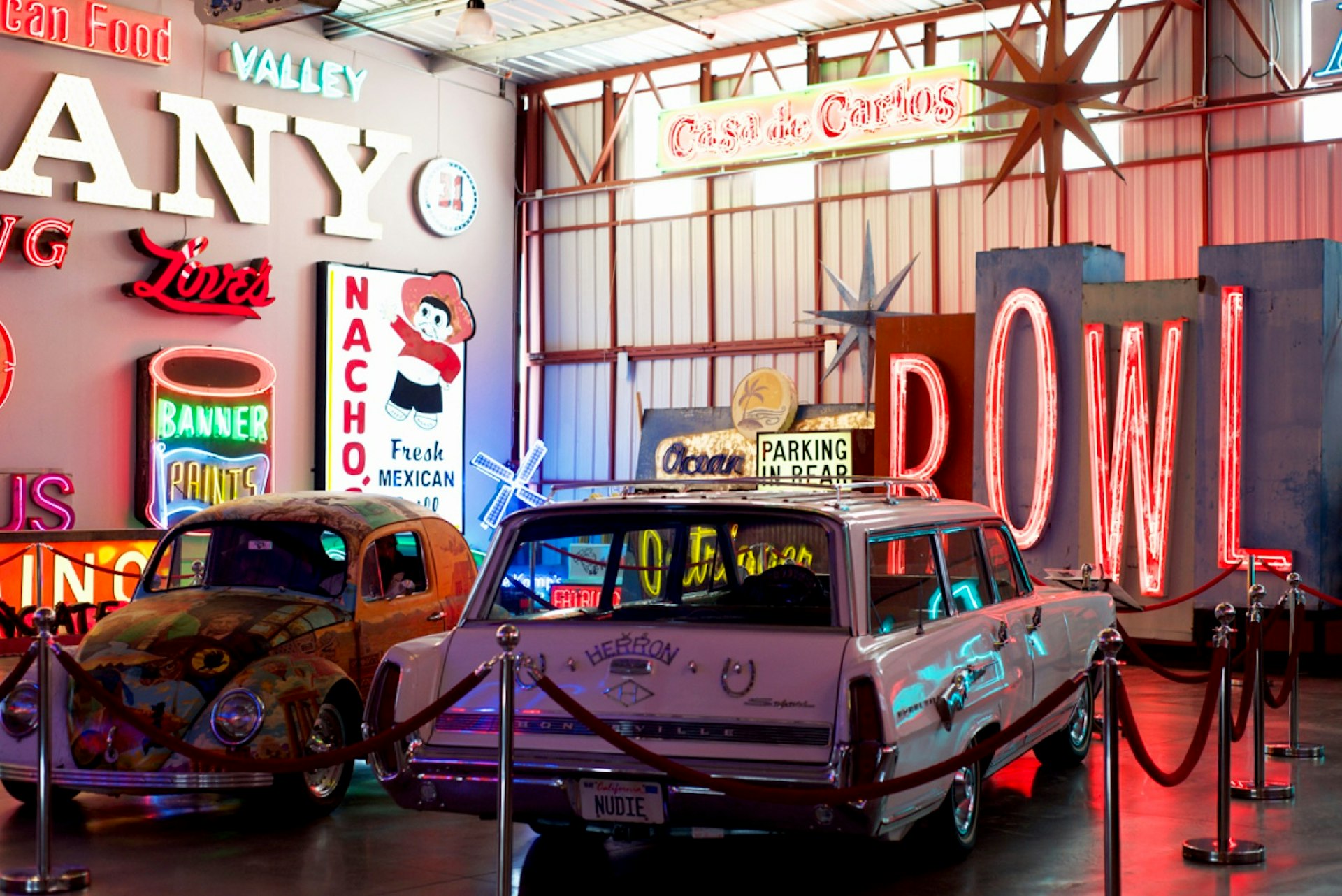 Several vintage signs, as well as decorated cars, are roped off in a warehouse-like exhibition; Los Angeles neon