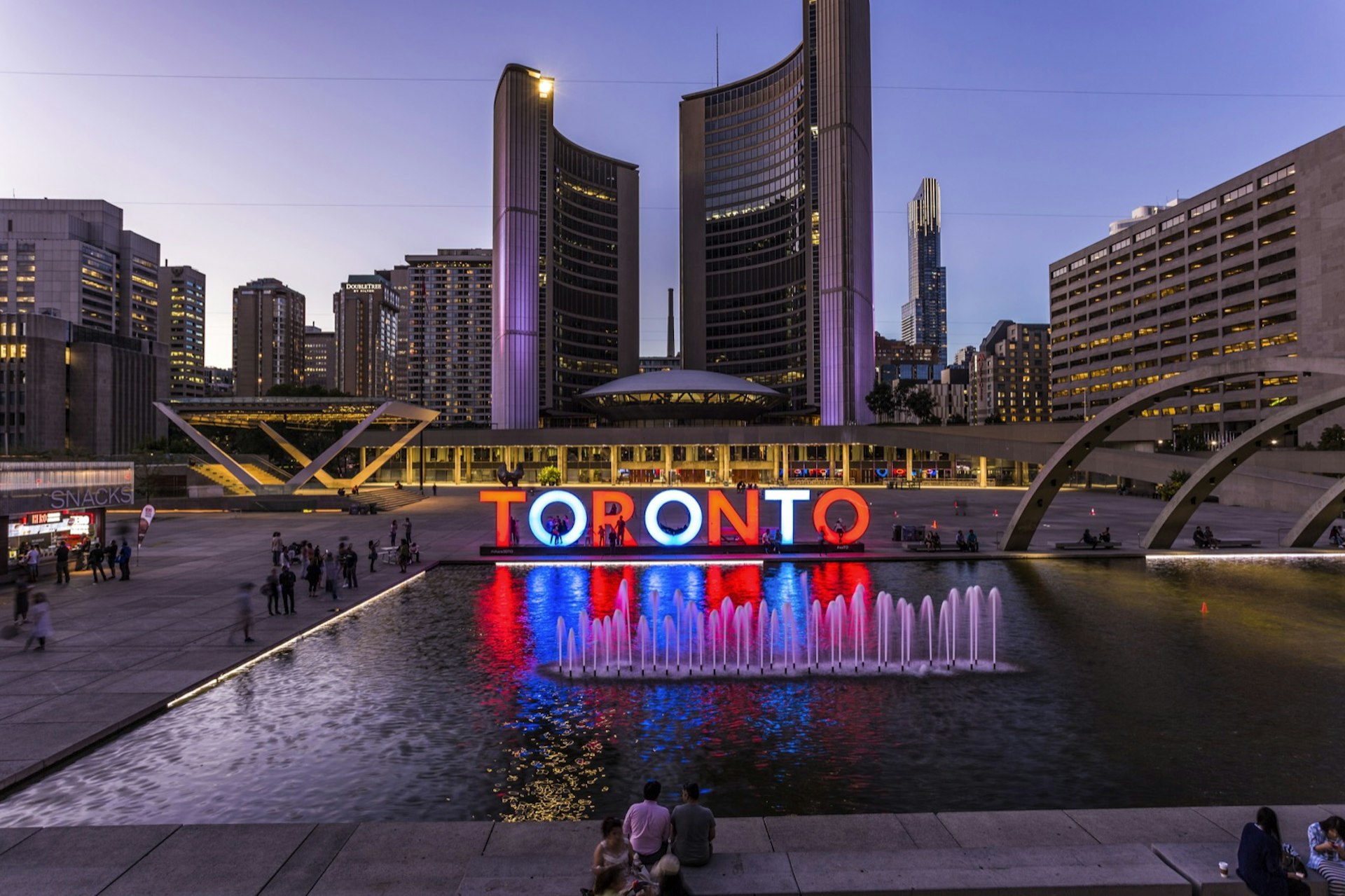 Torontoskylten vid Nathan Phillips Square lyser upp i rött och vitt och blått och reflekteras i en pool när solen går ner i Toronto;  Helg i Toronto
