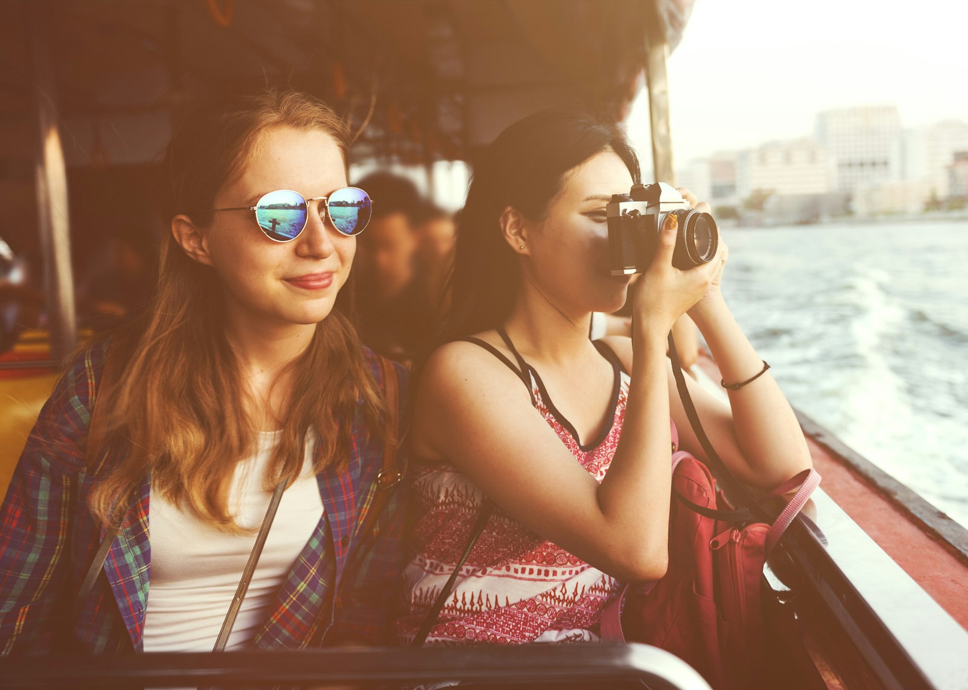 Two women sit together on a boat, looking out over the water. One is wearing sunglasses while the other looks through an SLR camera, pointing off the boat; travel and grief.
