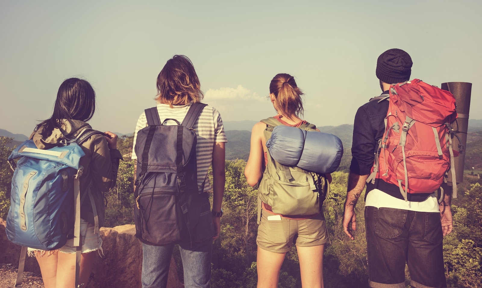 A shot of a group of four backpackers taken from behind. They all carry backpacks and have sleeping bags and various other items as they look out over a rural landscape; travel and grief 