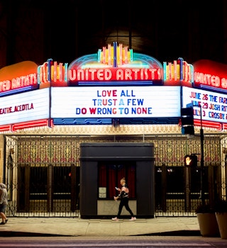 People walk in front of the Ace Hotel at night, which has an old-fashioned movie house marquee lit up in neon; Los Angeles neon